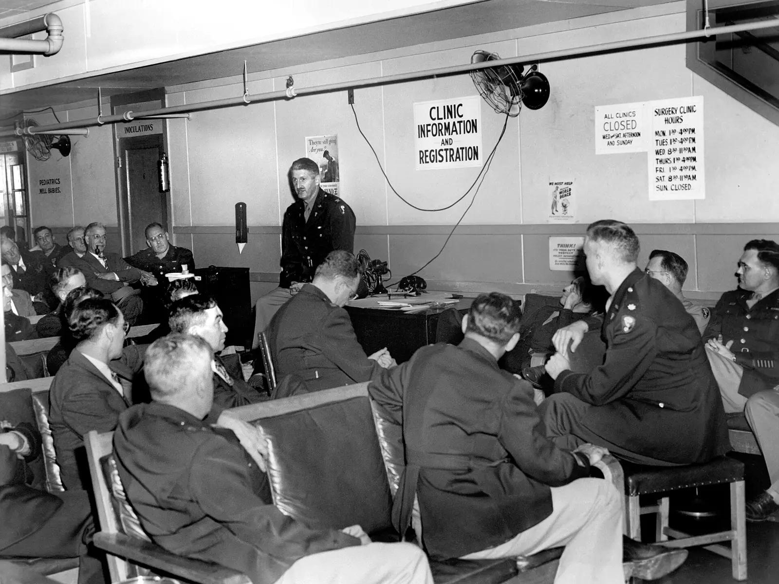 A staff meeting in a 1945 hospital waiting room, staff seated in rows facing the speaker